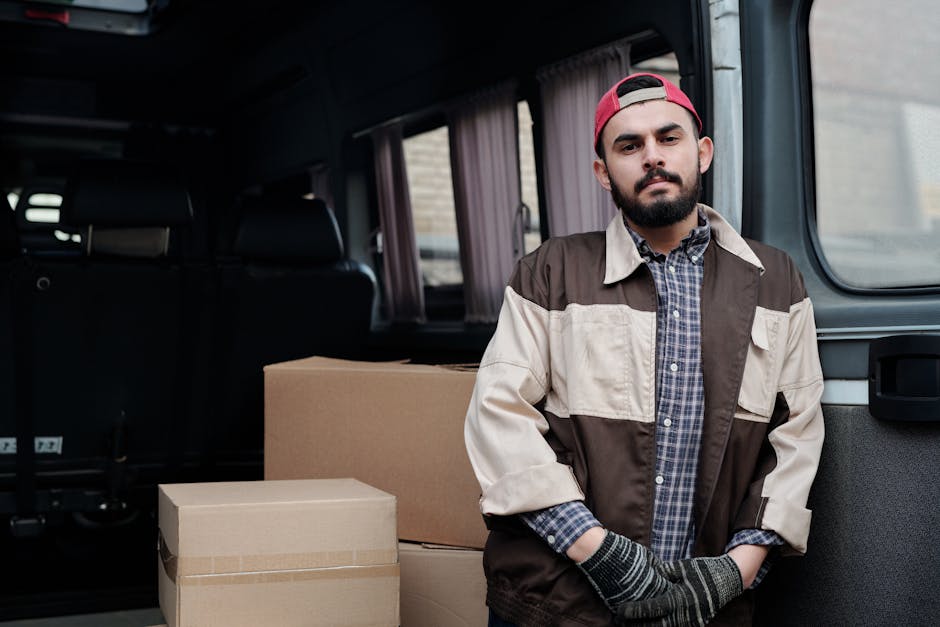 A man is standing beside a white moving van with its rear doors open, preparing for a home relocation. He is holding a cardboard box and appears to be in the process of loading or unloading. Several c