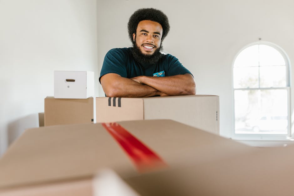 A young male mover with long, wavy hair and a black bandana is sitting in the driver’s seat of a white van branded with 'MOVING COMPANY LOCAL & LONG DISTANCE'. He is smiling and giving a thumbs-up t