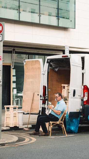 A man with glasses, wearing a light blue shirt and dark trousers, is seated on a wooden chair at the rear of a white van parked on a city street outside a modern building with glass windows and a beige facade. The van's side door is open, revealing a spacious interior loaded with large cardboard boxes, wooden furniture, and packed items covered with plastic wrapping and blankets, indicating a home relocation or furniture transport process. Nearby, a wooden pallet and a small wooden furniture piece are positioned on the pavement, and a step stool is placed next to the van for ease of loading. The scene captures the loading or unloading phase of a professional removals service, with visible equipment such as straps or blankets used to secure and protect furniture during transport, highlighting the logistical aspect of packing, moving, and house removals provided by Man with Van Brixton.