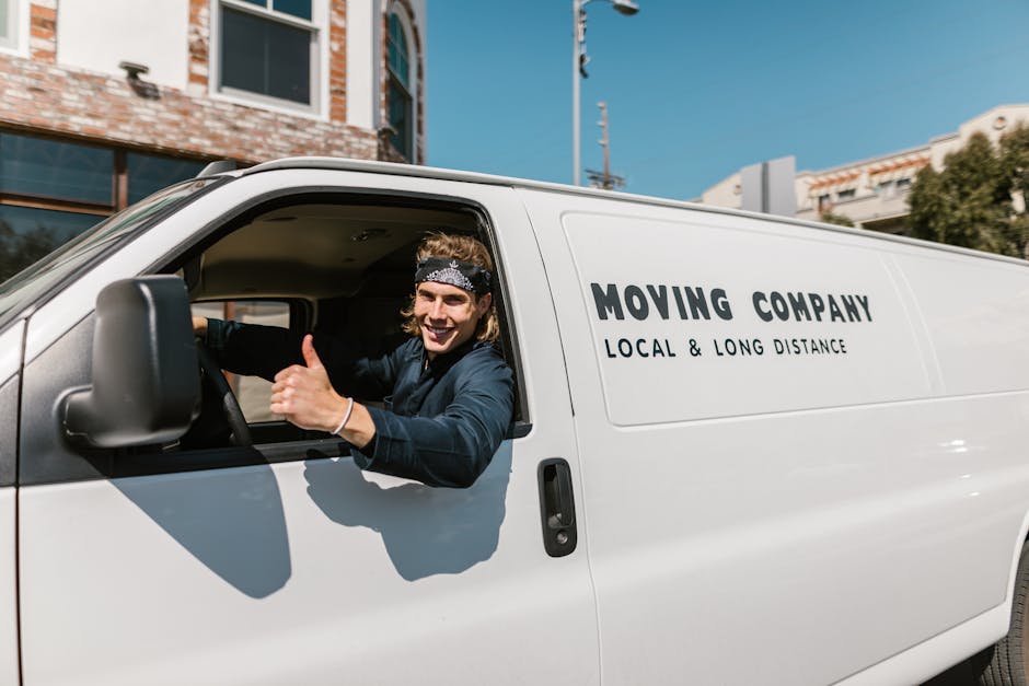 A smiling young man with long, wavy hair and a patterned headband is sitting in the driver’s seat of a white moving van parked outside a building, giving a thumbs-up gesture. The van, operated by Man with Van Brixton, features branding indicating they provide local and long-distance removals. The vehicle is positioned on a paved street with clear blue sky overhead, and in the background, there are modern buildings with large windows and some greenery. Inside the open driver’s side window, the man is visible wearing a dark long-sleeved shirt, and appears to be preparing for a house relocation or furniture transport task as part of a professional moving service. The scene captures the loading process or readiness for home removal, with the van's exterior clean and the man appearing confident and approachable for moving and packing services offered by [COMPANY_NAME], situated in Brixton.