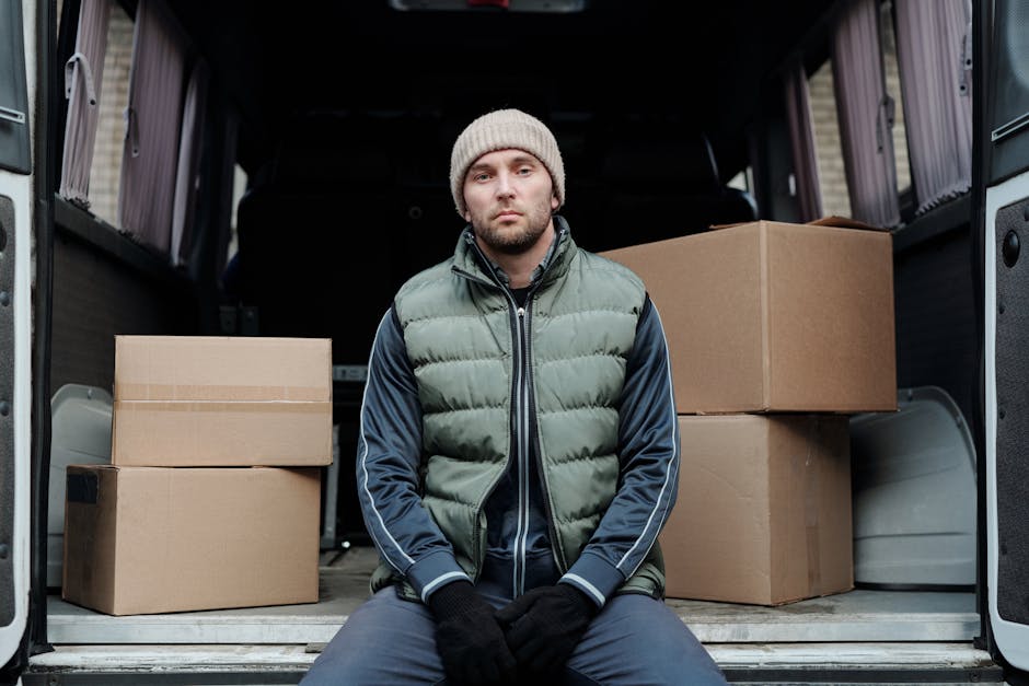 A man wearing a beige knit beanie, a green padded vest over a navy blue tracksuit, and black gloves is sitting on the ground in front of an open moving van. Behind him, the vehicle's interior shows several cardboard boxes stacked on the floor, some sealed with packing tape, and wrapped in plastic or fabric protective coverings. The boxes vary in size, with two larger ones on left and right and smaller boxes in between. The van's interior is illuminated with natural light, revealing the dark sides and ceiling. The scene depicts the process of loading or preparing for a home relocation, with the man seated on the pavement outdoors, possibly on a residential street or driveway, close to the building entrance. This image illustrates furniture transport and packing during a professional removal service, supporting the services offered by Man with Van Brixton, as indicated by the context of house removals in Brixton Market SW9.