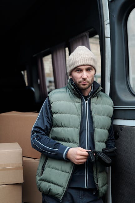 A man with a serious expression stands next to the open sliding door of a dark grey van, which is part of a home removal or furniture transport process. He is wearing a white knit beanie, a green padd